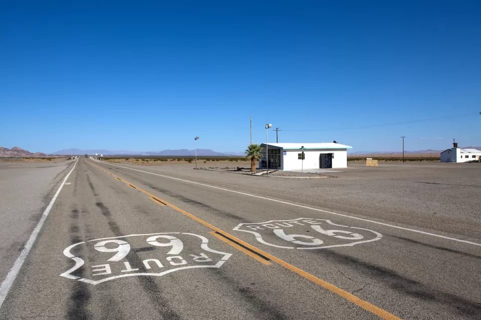 Low shot of Route 66 signs on asphalt with small abandoned building on roadside on old Route 66 near Amboy, California.